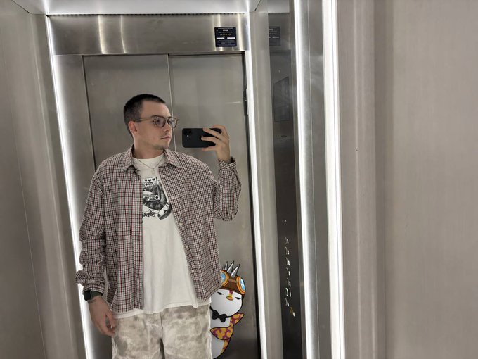 A mirror selfie of a man in sunglasses, a plaid shirt over a white t-shirt, and light pants, standing in an elevator next to a cartoon fox sticker.