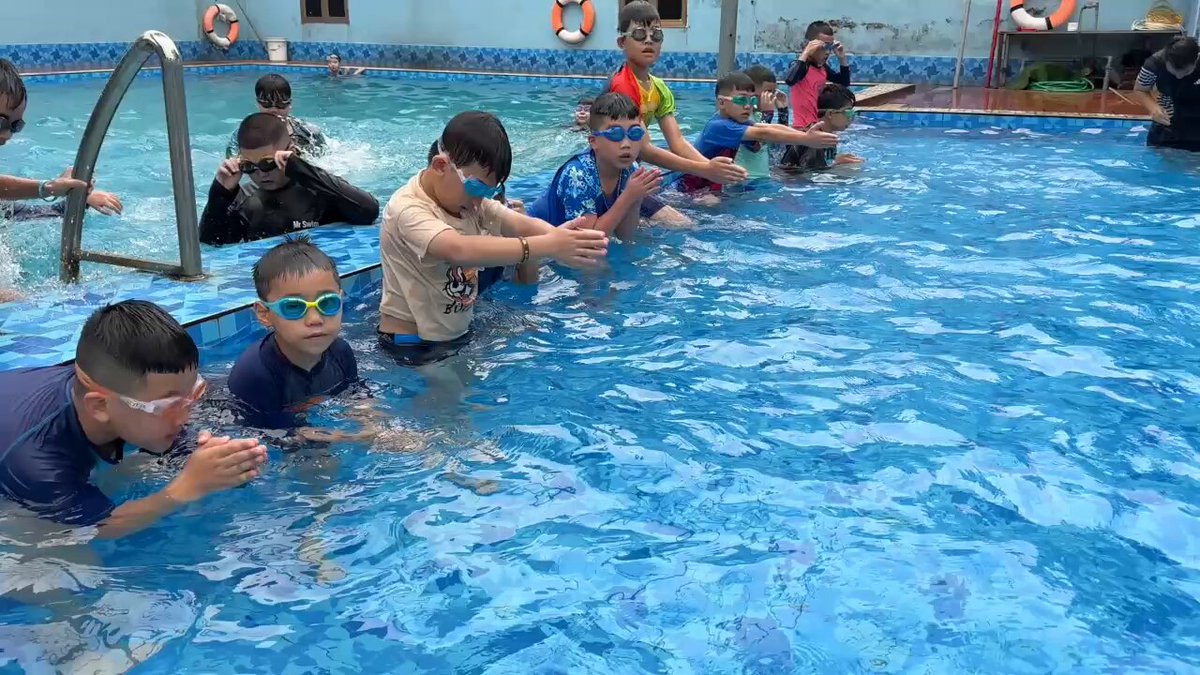 The image shows a group of children playing in a swimming pool. There are at least 12 people visible in the scene, with some kids sitting or standing near the edge of the pool while others are actively swimming. The children appear to be enjoying themselves as they interact and play together in the water.