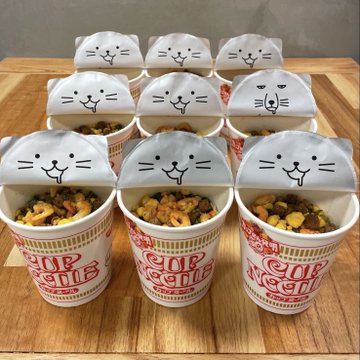 Multiple Cup Noodle containers arranged in rows with paper flaps decorated with cute hand-drawn cartoon faces popping up from the lids.