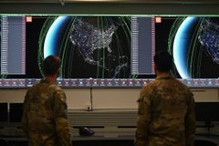 Two soldiers in camouflage uniforms viewed from behind, standing in a dark operations center looking at large screens displaying world maps with flight trajectories.