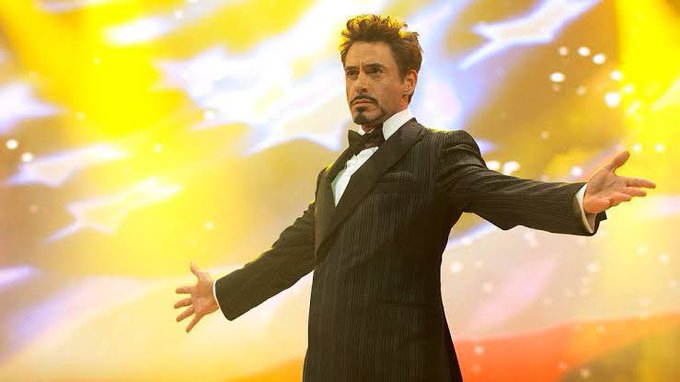 A man in a black tuxedo stands confidently with arms outstretched against a vibrant, explosive yellow background, symbolizing readiness and triumph. The post's motivational message about trying new things and grinding to win pairs perfectly with the image's heroic, open-armed pose suggesting an embrace of new challenges and impending success.