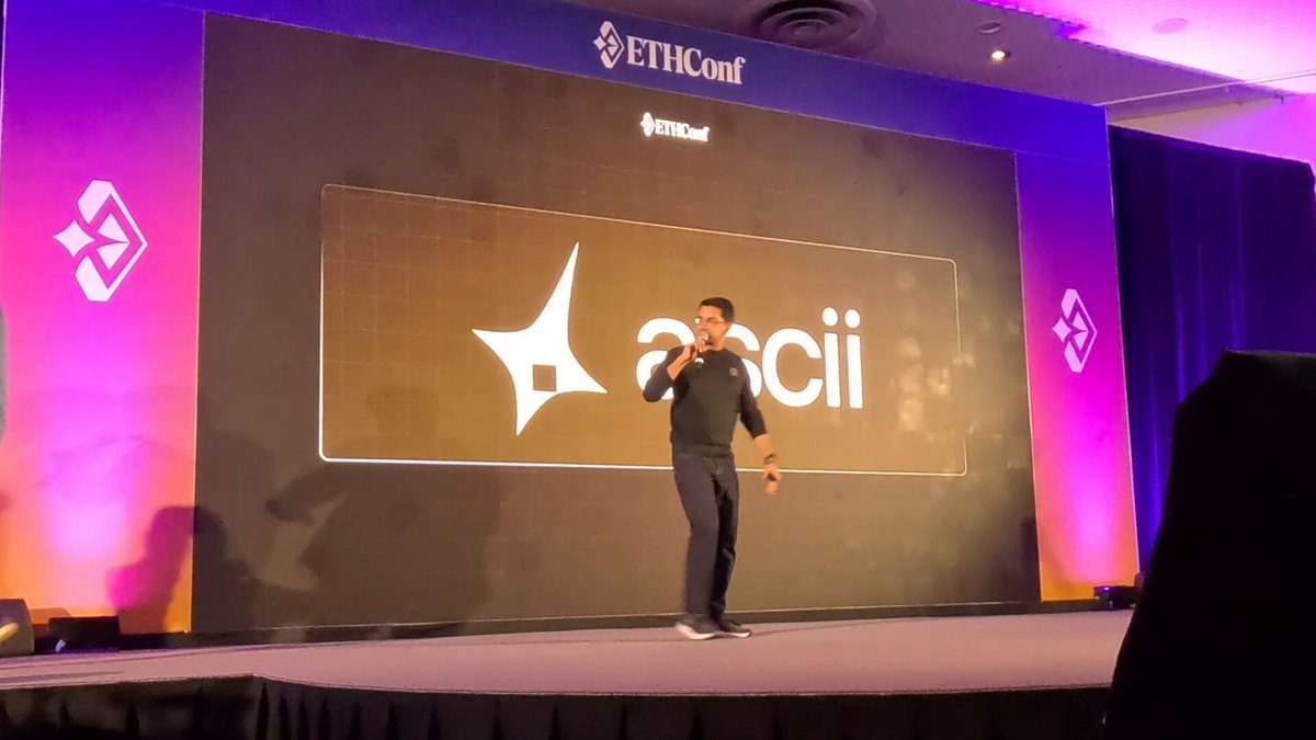 The image shows a man standing on a stage, giving a speech or presentation. He appears to be wearing a black shirt and pants. There are two people visible in the background of the scene, one near the left side and another person closer to the center. The stage is set up with purple lights behind the speaker, creating an atmosphere for his presentation. A banner can also be seen on the right side of the image, possibly displaying information related to the event or the speaker's name.