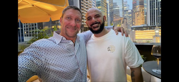 Two men smiling and posing together for a photo on an outdoor patio at dusk, with a city skyline and yellow umbrellas visible in the background.