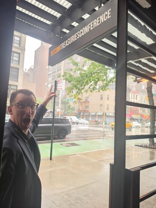 A man in glasses and a suit excitedly points to a sign reading 'BITCOINTREASURIESCONFERENCE' while standing under a covered walkway on a rainy city street.