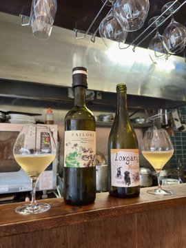 The image features two wine bottles placed next to each other on a wooden countertop or bar. The bottles are filled with wine, one of which appears to be a red wine. There are also two wine glasses positioned near the bottles, ready for serving. In addition to these elements, there is an oven in the background and a microwave above it. A cell phone can also be seen on the countertop.