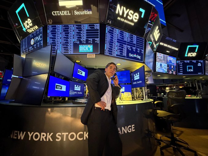 A man in a suit talks on a phone at the New York Stock Exchange trading floor, surrounded by digital displays showing "NYSE," "ICE," "Citadel Securities," "Dick's," and various ticker boards. The "TOKENISE IT ALL" caption ties to the image's depiction of the NYSE trading environment—the iconic traditional finance hub where tokenization efforts aim to digitize and revolutionize asset trading.