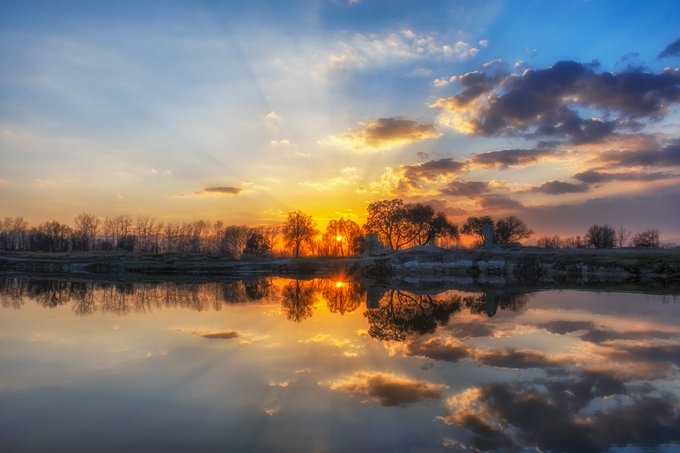 A scenic sunset landscape with golden and orange clouds reflecting perfectly in a calm body of water, with silhouettes of trees along the horizon.