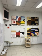 A photo of a room with white walls and a tiled floor, displaying two Mondrian-style geometric color block compositions taped to the wall above a wooden cart.
