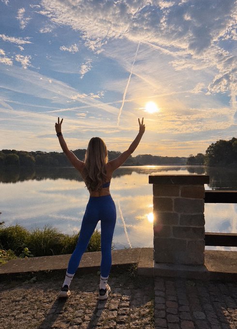 A woman in a blue athletic outfit stands with her back to the camera, raising both arms in peace signs towards a sunset over a calm lake.