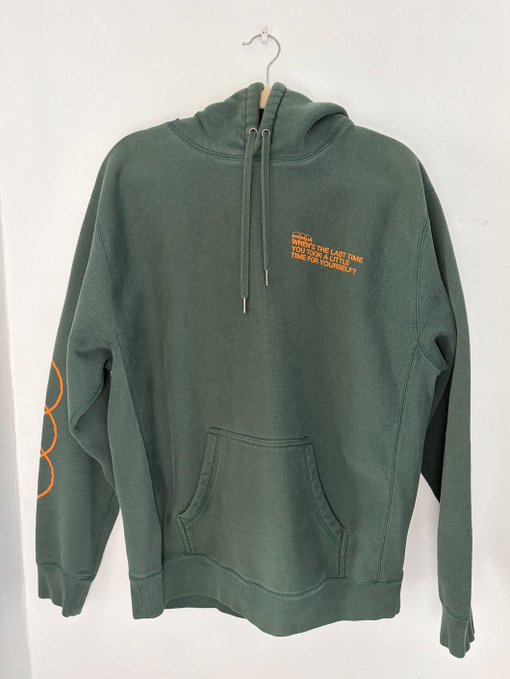 A faded sage green pullover hoodie hangs on a white hanger against a plain wall, featuring orange text on the chest reading "STREETS THE LAST TIME YOU SAW A LITTLE TIME FOR YOUR TOMORROW" with an orange circular graphic on the left sleeve—fitting the casual "favorite hoodie" reply while subtly inviting the promised airdrop.