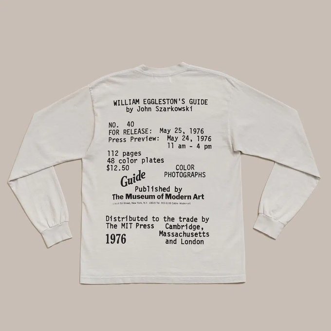 A white long-sleeve t-shirt displays text on the back promoting 'William Eggleston's Guide by John Szarkowski', published May 25, 1976 by The Museum of Modern Art and distributed by The MIT Press.