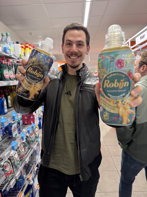 A man in a black leather jacket and olive shirt smiles while holding up two bottles of "Robijn" fabric softener—one "Creations Camelot Vanilla" and one "Creations Pareo Brezza"—in a supermarket aisle, humorously riffing on his post's "laundry detergent content" joke. The playful product endorsement visually underscores his self-deprecating comparison of the current bearish market sentiment to being stuck in a wash cycle of bad news.