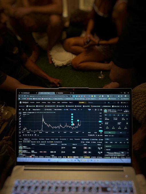 A laptop displays a cryptocurrency trading chart on what appears to be the Trojan exchange, with people sitting on green carpet in the background.