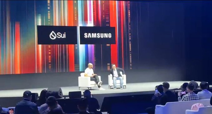 Two men sit in white armchairs on a stage in front of a large screen displaying the Sui and Samsung logos, with an audience visible in the foreground.