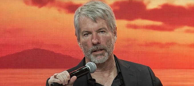 A close-up of a gray-haired, bearded man in a black suit holding a microphone while speaking on stage with a red and orange sunset backdrop.