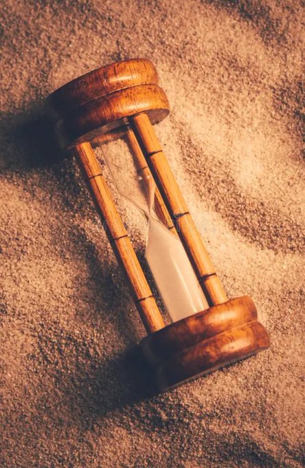 An antique wooden hourglass with white sand resting on a sandy surface, casting a warm shadow.