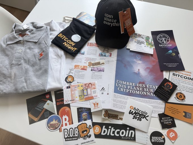 A collection of Bitcoin and cryptocurrency-themed merchandise is displayed on a white table, including a black 'open source everything' cap, a gray polo shirt, a white t-shirt, a 'BITCOIN NIGHT' bag, and various stickers and flyers.