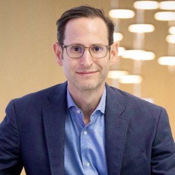 Matthew Sigel, head of digital assets research at VanEck, is shown in a professional headshot wearing glasses and a blue blazer over a light blue shirt, with a warmly lit interior background. The image accompanies his bullish outlook on Bitcoin's potential to reach $100,000 next year and $500,000 by 2029, lending a face to VanEck's optimistic crypto forecast.