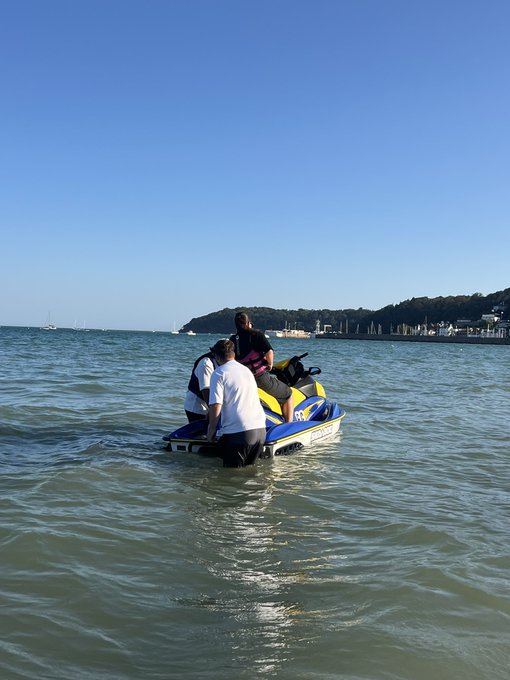 Two people stand in shallow turquoise water behind a blue and yellow jet ski, with a distant shoreline and sailboats visible under a clear blue sky. The casual, sunny scene reflects a laid-back "Good Morning" vibe, fitting the post's invitation to drop a $SOL crypto address.