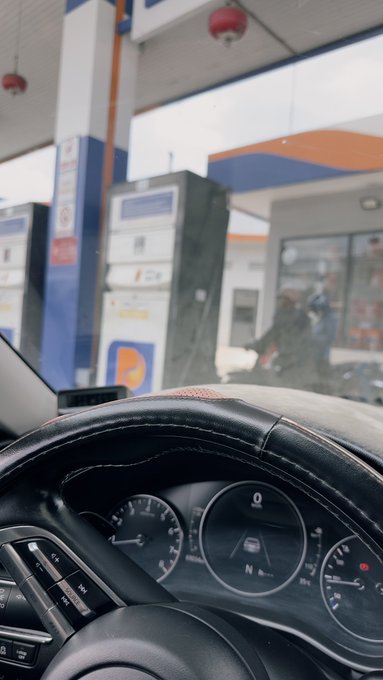 A driver grips the steering wheel of a car at a gas station, with the dashboard showing the gear in "N" (Neutral) and the fuel gauge appearing nearly empty. The pump price is visible at over 24,600 VND per liter, reflecting the post's complaint about high fuel costs making it too expensive to fill up the tank.