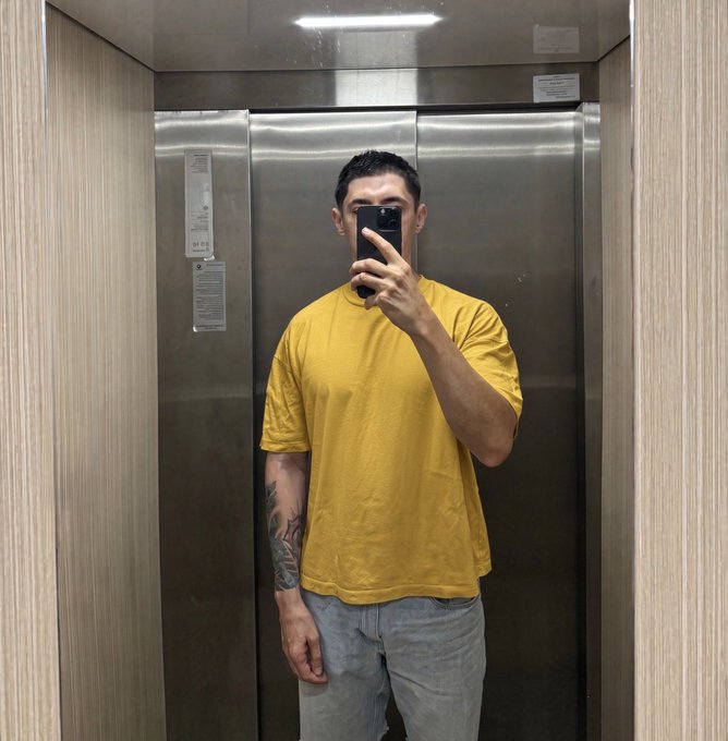 A man in a yellow t-shirt and light blue jeans takes a mirror selfie with his phone inside an elevator, showing a tattoo on his right arm.
