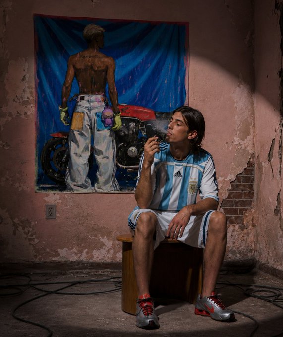 A man in an Argentina soccer jersey sits smoking in front of a painting depicting a tattooed figure beside a motorcycle, with cables strewn across the floor—matching the post's promotion of the artwork "Sunset of the Gypsy" available as a 1/1 on Tezos.