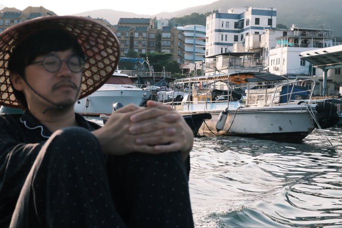 A man wearing a wide-brimmed conical hat and glasses sits with his hands clasped, gazing contemplatively across a harbor filled with fishing boats and waterfront buildings. The reflective, pensive mood of the scene visually echoes the post's meditation on legacy and the moral facades historically worn by profiteers.