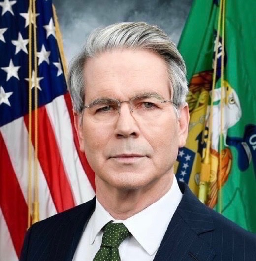 An official portrait of a gray-haired man wearing glasses, a dark suit, and a green tie, positioned in front of the United States flag and the green flag of the U.S. Department of the Treasury.
