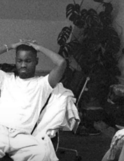 A black-and-white photo of a man in a white t-shirt with his hands behind his head, seated indoors with a plant visible in the background.