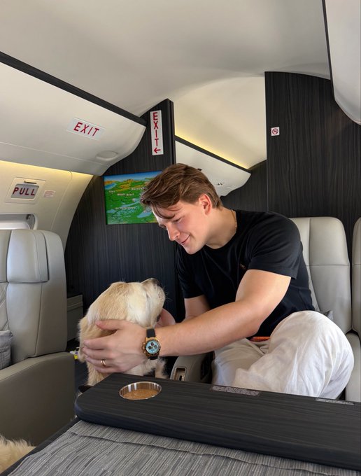 A man sits in the cabin of a private jet, smiling as he pets a fluffy white dog, with 'EXIT' signs visible above and a moving map displayed on a screen.