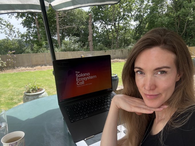 The image features a woman sitting at an outdoor table, holding her chin up as she poses for a photo. On the table, there's a laptop open with the words "Solana Ecosystem Call" displayed on its screen. The setting appears to be a sunny day in a park or garden area, and there is an umbrella providing shade nearby.