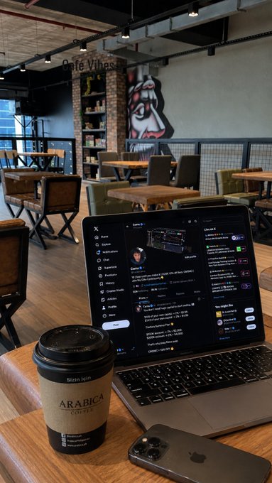 A laptop on a wooden table at 'Café Vinho' displays an X (formerly Twitter) feed, with an 'ARABICA COFFEE' cup and a smartphone beside it.