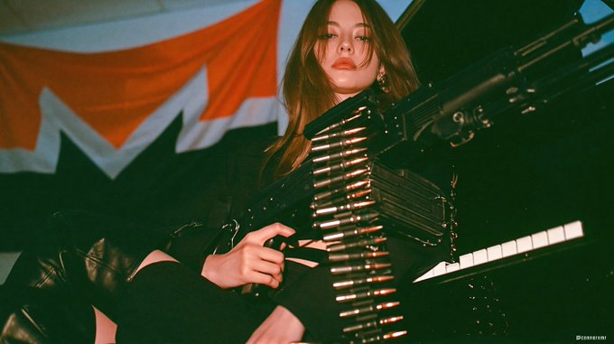 A young woman holds a large firearm with an ammunition belt draped across it, with a red and white flag in the background.