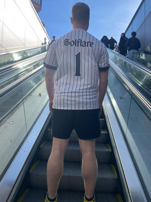 Person seen from behind riding an escalator wearing a striped baseball-style jersey that reads "Solflare 1" on the back, with black shorts.