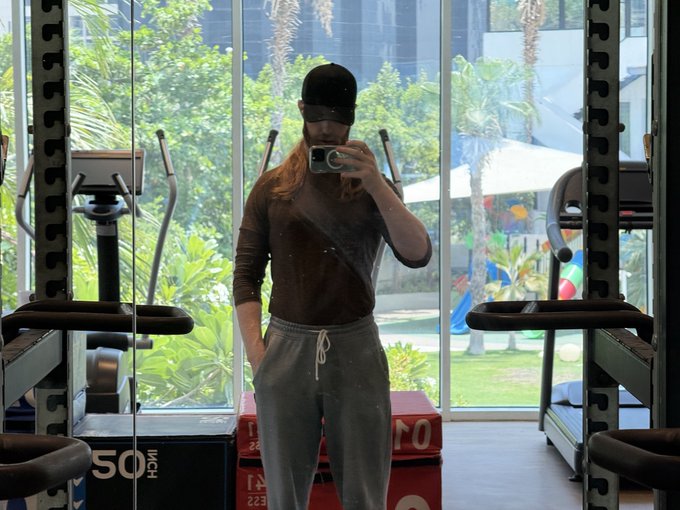 A person wearing a black cap, sunglasses, brown long-sleeve shirt, and gray sweatpants takes a mirror selfie with their phone in a gym, with treadmills and a tropical outdoor view visible in the background.