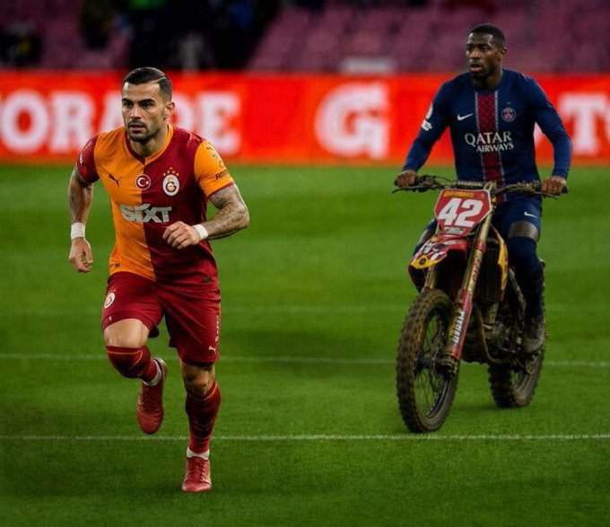 A soccer player in a Galatasaray jersey runs on a field while a person on a dirt bike rides alongside, featuring "QATAR AIRWAYS" on the jersey.