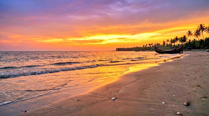 The image shows a beach scene with a boat floating near the shoreline, possibly docked at a pier or moored to a dock. The sky above has an orange hue, indicating that the sun is setting over the ocean. The water appears calm, suggesting a peaceful and serene atmosphere.