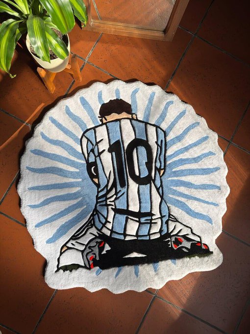 An Argentina soccer jersey-shaped rug featuring the number 10 and Messi kneeling, placed on a tiled floor next to a potted plant.