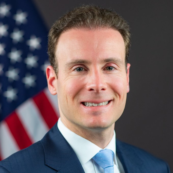 An official portrait of a smiling man in a blue suit and light blue tie, standing in front of an American flag.