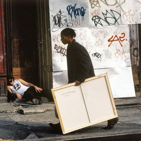 A candid street photograph showing a man carrying a large blank canvas while another man lies on the sidewalk near a graffiti-covered doorway at what appears to be an urban gallery or art location.