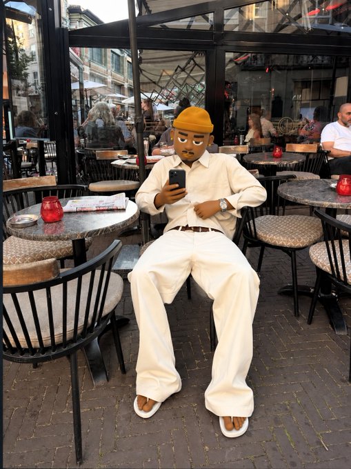 A person dressed in cream-colored clothing and an orange beanie sits at an outdoor café table looking at their phone, with a cartoon face emoji covering their face and other patrons visible in the background.