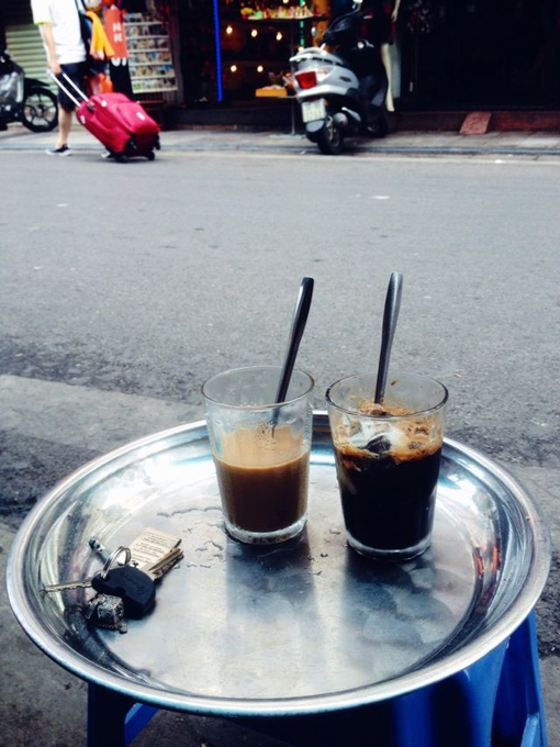Two glasses of iced coffee sit on a small metal tray on a street sidewalk, with a red suitcase and motorbike visible in the background.
