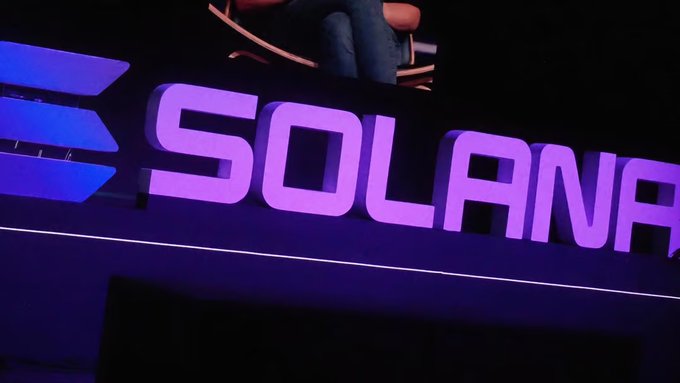 The image shows an illuminated "SOLANA" sign in purple and blue lighting at what appears to be an event venue, with the brand name prominently displayed against a dark background. The image ties to the post about Solana's governance vote on token issuance reduction, visually representing the blockchain network at the center of the onchain voting discussion.