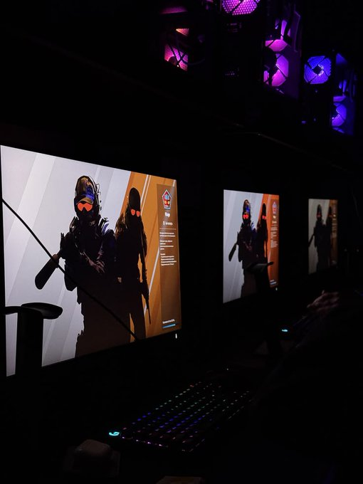 A dimly lit photograph of a gaming setup featuring an RGB illuminated keyboard and three monitors displaying video game characters holding weapons, with PC components lit in purple above.