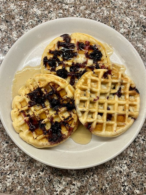 Three homemade waffles topped with blueberry compote and syrup are served on a white plate sitting on a granite countertop.
