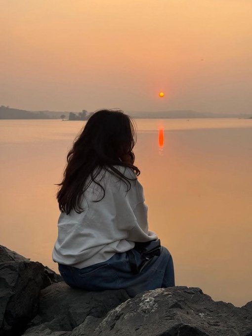 A woman with long dark hair sits on rocky terrain, gazing out at a calm body of water as the sun sets, casting an orange glow across the horizon.
