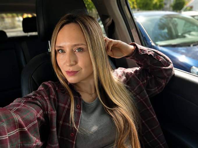 A woman with long blonde hair sits inside a vehicle, looking toward the camera with one hand near her head, wearing a maroon and green plaid flannel over a gray top. The casual selfie pairs with a warm Friday greeting, suggesting a personal "good morning" check-in from a Web3 influencer to their community on Crypto Twitter.