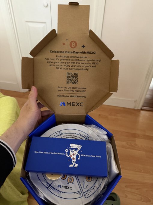 The image shows a person holding an open pizza box with a blue lid that has a white logo on it. The box contains a pizza cutter, a wallet, and a coin inside. There are also two coins visible in the background of the image.