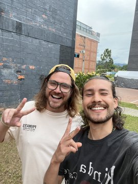 Two smiling young men pose outdoors in front of a modern gray stone building; one wears a yellow cap and glasses and flashes peace signs, while the other sports long hair and a black shirt.