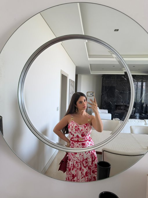 A woman in a red and white patterned dress takes a mirror selfie in a modern interior with a large round mirror.